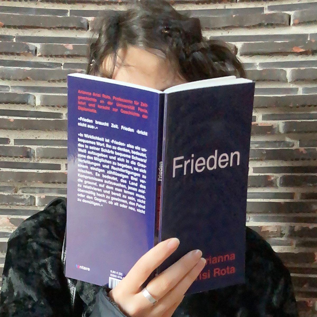 Arianna Arisi Rota: Frieden, tentare Verlag – aufgeschlagenes dunkelblaues Taschenbuch, das eine lesende Person vor ihr Gesicht hält, vor einer Wand aus dunklen Klinkersteinen
