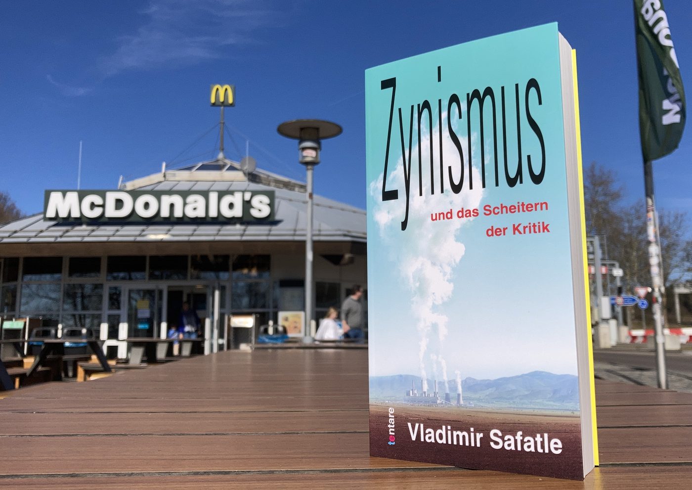 Vladimir Safatle: Zynismus und das Scheitern der Kritik, tentare Verlag – Buch mit Cover (Kraftwerk mit Dampfwolke vor blauem Himmel) auf einem Holztisch vor einem McDonald’s-Schnellrestaurant