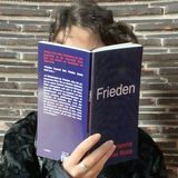 Arianna Arisi Rota: Frieden, tentare Verlag – aufgeschlagenes dunkelblaues Taschenbuch, das eine lesende Person vor ihr Gesicht hält, vor einer Wand aus dunklen Klinkersteinen